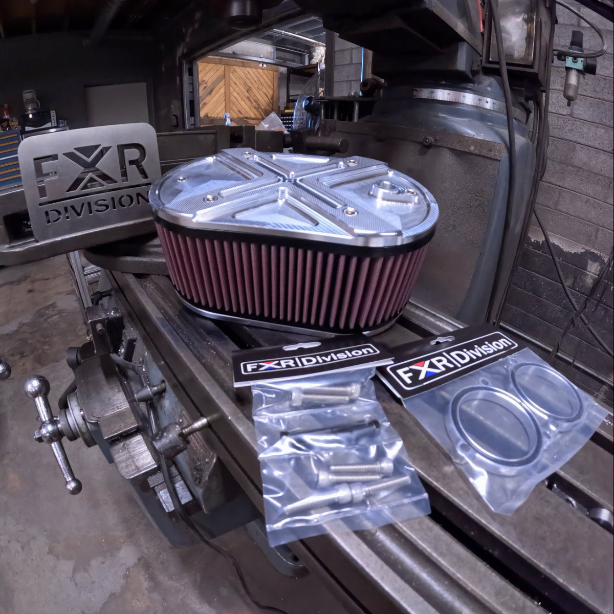 An FXR Division / TPJ Concepts Hi-FLOW PERFORMANCE AIR CLEANER for RevMax models, shown with mounting hardware, air filter, and CNC machined aluminum body, displayed on a workbench.